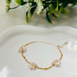 May include: A delicate gold chain bracelet with three small pearl flower charms. The bracelet is on a white surface.