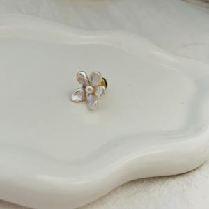 May include: A delicate flower-shaped pin with a pearl center and petal-like shapes. The pin is made of a light-colored material, possibly pearl or shell, and has a gold-colored pin backing. The pin is set against a white background.