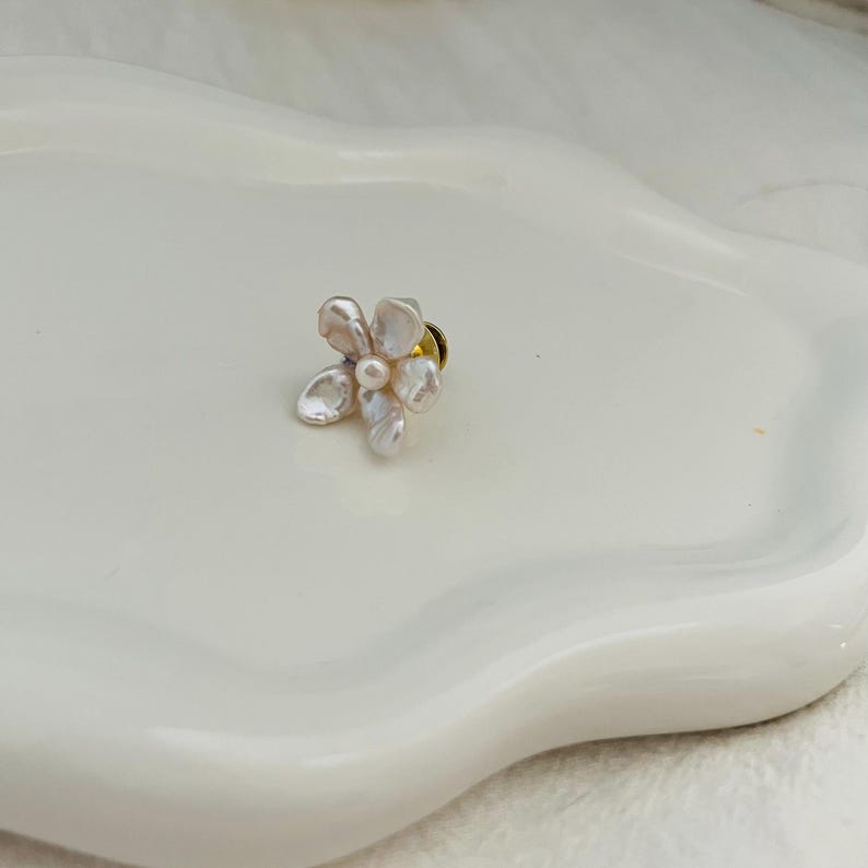 May include: A delicate flower-shaped pin with a pearl center and petal-like shapes. The pin is made of a light-colored material, possibly pearl or shell, and has a gold-colored pin backing. The pin is set against a white background.
