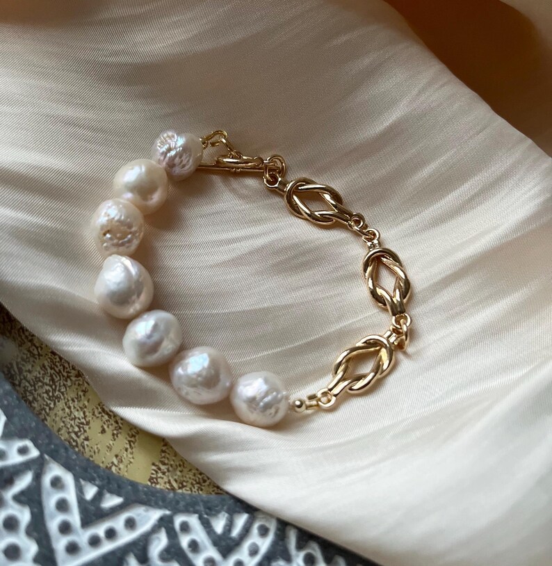 Puede incluir: Un brazalete de cadena dorada con un dise&ntilde;o anudado y una fila de perlas blancas.