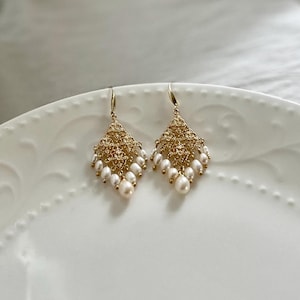 Peut inclure: Une paire de boucles d'oreilles pendantes dor&eacute;es avec une structure en m&eacute;tal ajour&eacute; en forme de losange. Chaque boucle est orn&eacute;e de plusieurs petites perles d'eau douce blanches en forme de goutte suspendues au bord inf&eacute;rieur.