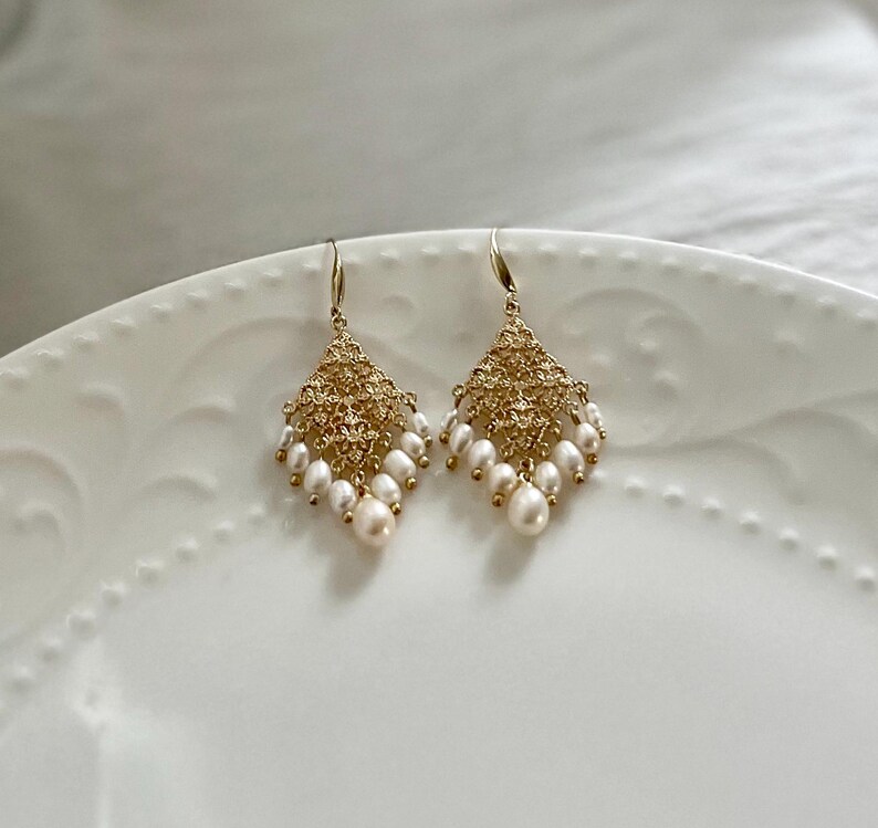 Peut inclure: Une paire de boucles d'oreilles pendantes dor&eacute;es avec une structure en m&eacute;tal ajour&eacute; en forme de losange. Chaque boucle est orn&eacute;e de plusieurs petites perles d'eau douce blanches en forme de goutte suspendues au bord inf&eacute;rieur.