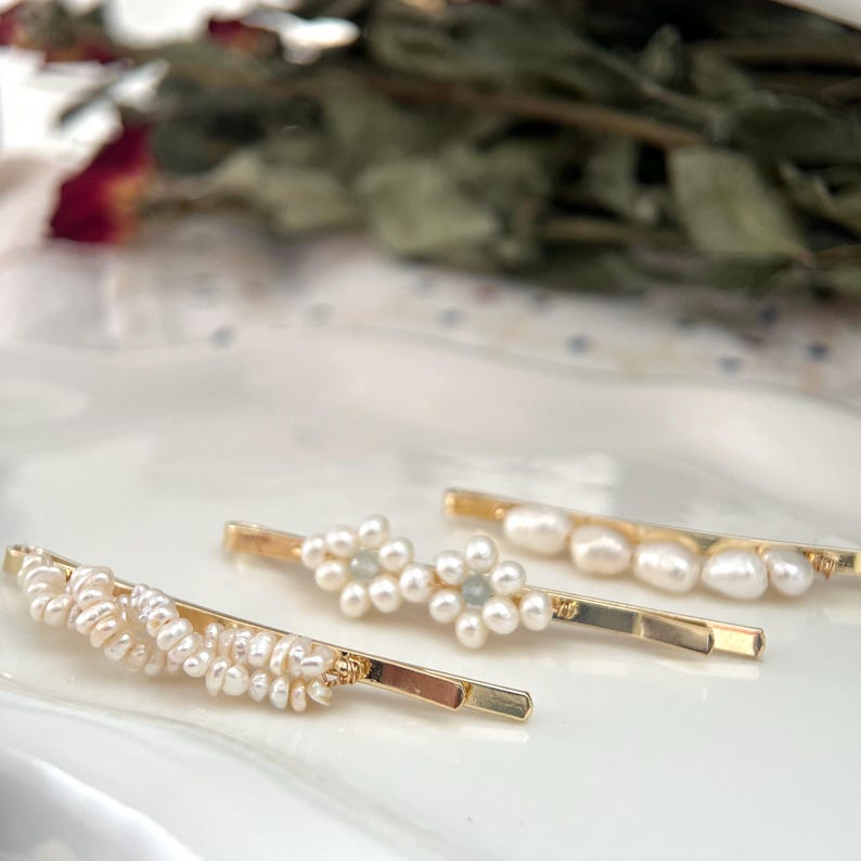 Puede incluir: Cuatro horquillas doradas para el cabello con adornos de perlas y cuentas. Una horquilla presenta un grupo de peque&ntilde;as perlas, mientras que otra tiene un dise&ntilde;o floral con perlas y peque&ntilde;as cuentas azules. Las otras dos tienen detalles de perlas.