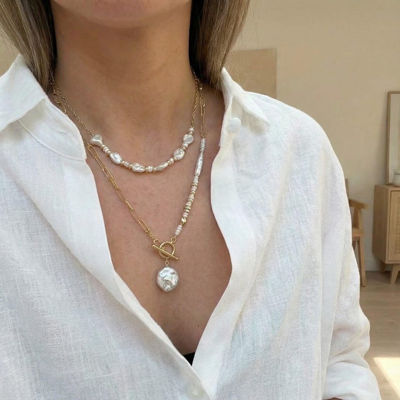 May include: Two-strand gold-tone necklace set. The top strand features irregular white pearls, while the lower strand has a large, baroque pearl pendant. The necklace is worn over a white button-down shirt.