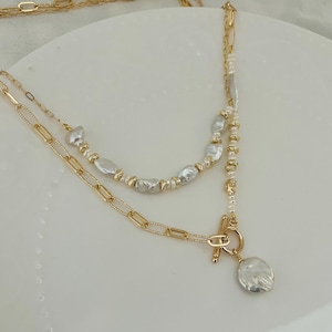 May include: A layered gold-coloured necklace with a toggle clasp and a large, irregular-shaped pearl pendant. The necklace features a chain link design and is adorned with smaller pearls and gold beads.
