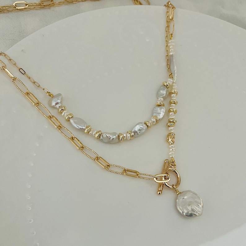 May include: A layered gold-coloured necklace with a toggle clasp and a large, irregular-shaped pearl pendant. The necklace features a chain link design and is adorned with smaller pearls and gold beads.