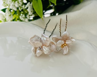Puede incluir: Dos horquillas florales con pétalos perlados y tallos metálicos. Una horquilla tiene un grupo de pequeñas cuentas plateadas, y la otra tiene un grupo de cuentas doradas. Las horquillas están colocadas sobre una superficie blanca con vegetación de fondo.
