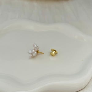 May include: A single floral earring with a gold post and a separate gold earring back. The earring features a cluster of white, petal-shaped elements. The earring is set against a white, wavy surface.