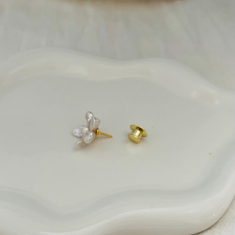 May include: A single floral earring with a gold post and a separate gold earring back. The earring features a cluster of white, petal-shaped elements. The earring is set against a white, wavy surface.