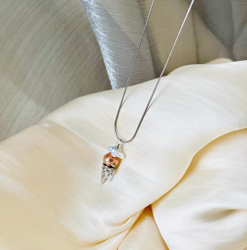 Peut inclure: Un collier en cha&icirc;ne argent avec un pendentif en forme de cornet de glace. Le cornet est en argent et a trois boules de fausses perles dans des tons de blanc et de p&ecirc;che.