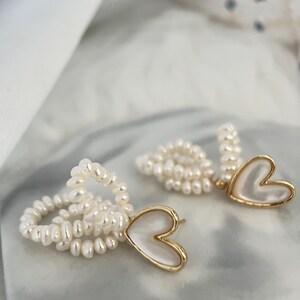 May include: A pair of gold earrings with a heart-shaped pendant made of white mother-of-pearl. The earrings are made of small white pearls.