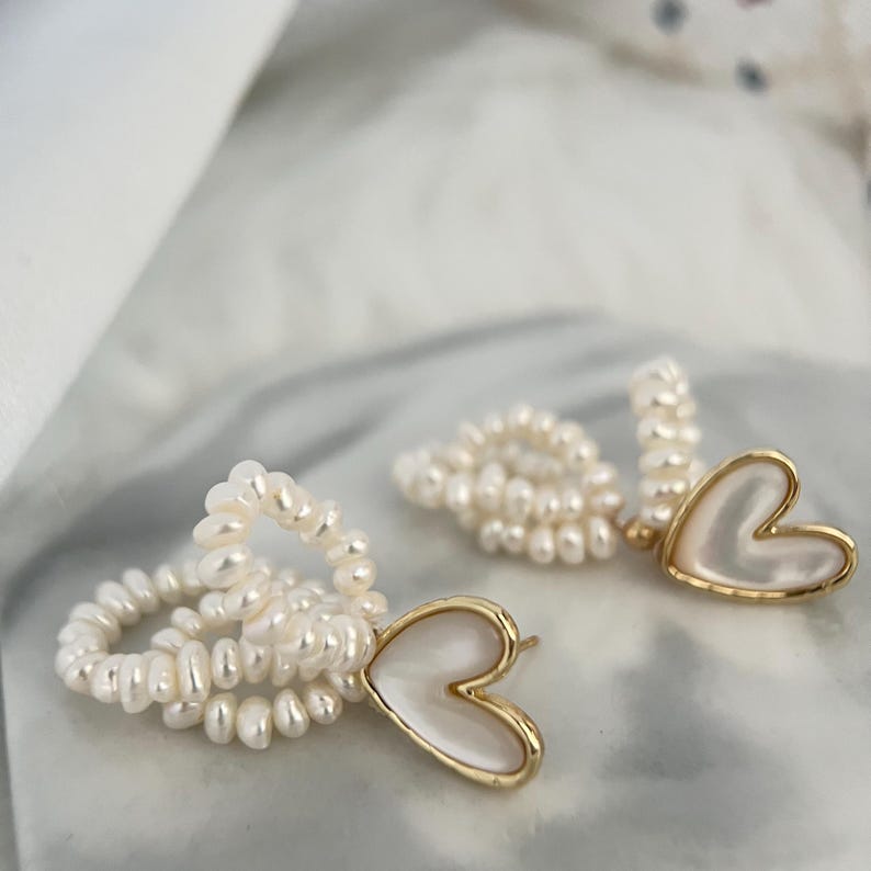 May include: A pair of gold earrings with a heart-shaped pendant made of white mother-of-pearl. The earrings are made of small white pearls.