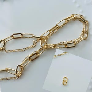 May include: Gold-coloured chain necklace featuring a variety of link shapes, including oval and rectangular. The necklace is displayed on a white surface. A small, gold-coloured clasp is also shown.