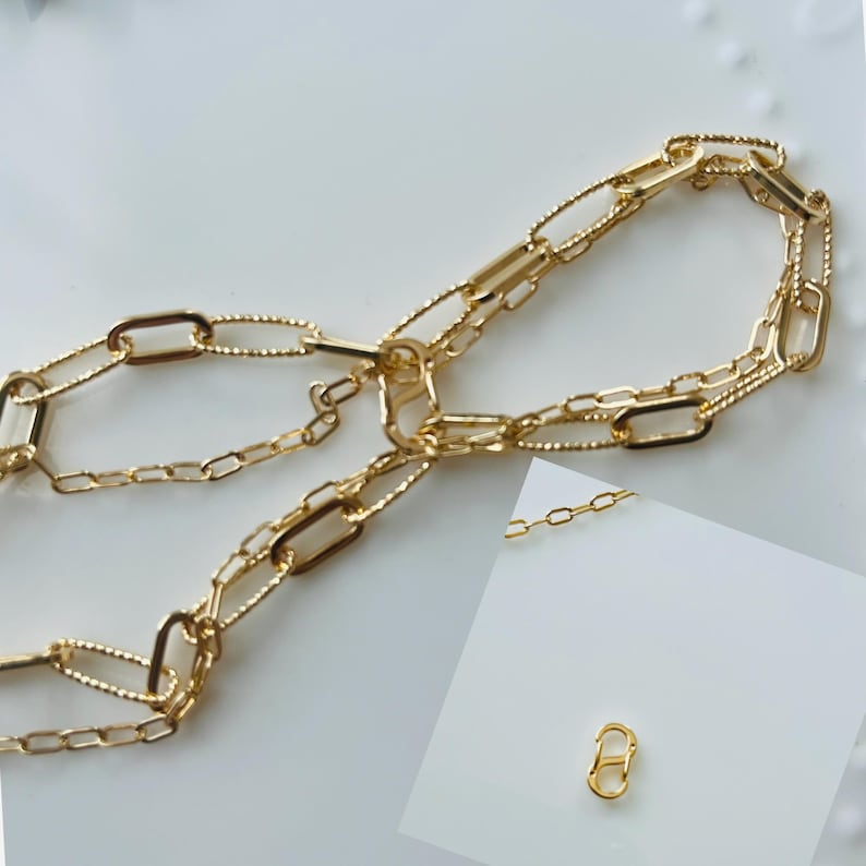 May include: Gold-coloured chain necklace featuring a variety of link shapes, including oval and rectangular. The necklace is displayed on a white surface. A small, gold-coloured clasp is also shown.