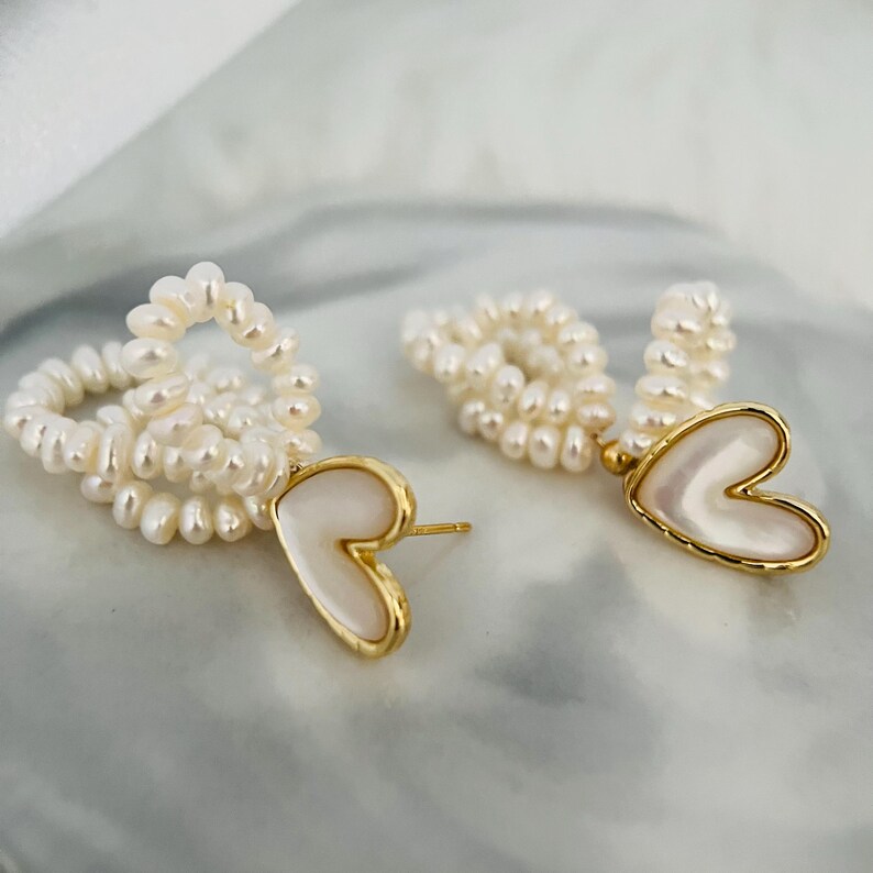 May include: A pair of gold earrings with a heart-shaped pendant made of white mother of pearl. The earrings are made of small white pearls.