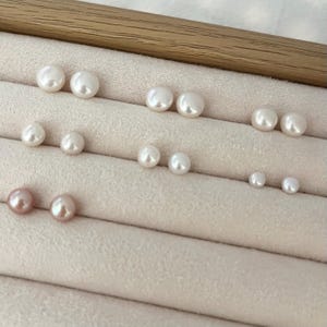 Peut inclure: Une collection de boucles d'oreilles en perles blanches et roses de diff&eacute;rentes tailles, dispos&eacute;es sur une surface beige clair. Les perles sont rondes et ont un &eacute;clat brillant, refl&eacute;tant la lumi&egrave;re. Les boucles d'oreilles sont pr&eacute;sent&eacute;es par paires.