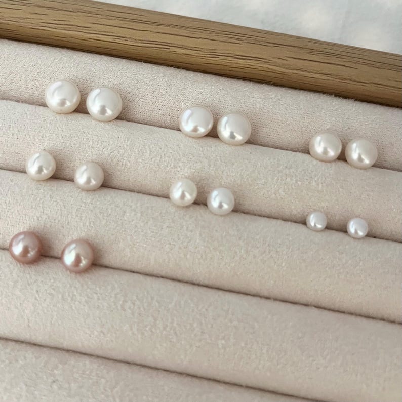 Peut inclure: Une collection de boucles d'oreilles en perles blanches et roses de diff&eacute;rentes tailles, dispos&eacute;es sur une surface beige clair. Les perles sont rondes et ont un &eacute;clat brillant, refl&eacute;tant la lumi&egrave;re. Les boucles d'oreilles sont pr&eacute;sent&eacute;es par paires.