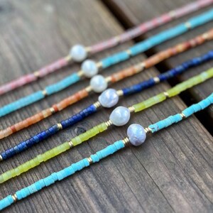May include: A set of six delicate necklaces featuring a variety of colorful gemstone beads and a single pearl accent. The necklaces are arranged in a rainbow pattern, with the colors ranging from pink to turquoise.