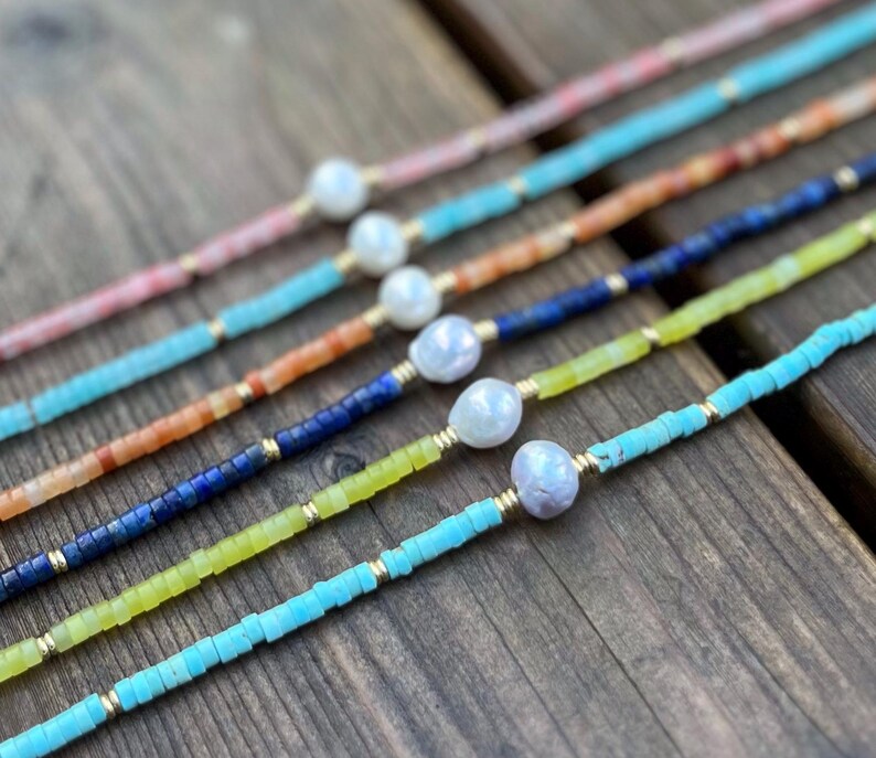 May include: A set of six delicate necklaces featuring a variety of colorful gemstone beads and a single pearl accent. The necklaces are arranged in a rainbow pattern, with the colors ranging from pink to turquoise.