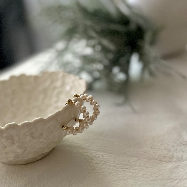 May include: A pair of gold hoop earrings with white pearl accents. The earrings are sitting in a white ceramic bowl with a floral design.