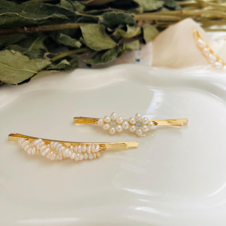 Puede incluir: Dos horquillas doradas. Una presenta un grupo de peque&ntilde;as perlas blancas, la otra un dise&ntilde;o floral de perlas. Se colocan sobre un fondo blanco, con vegetaci&oacute;n en el fondo.