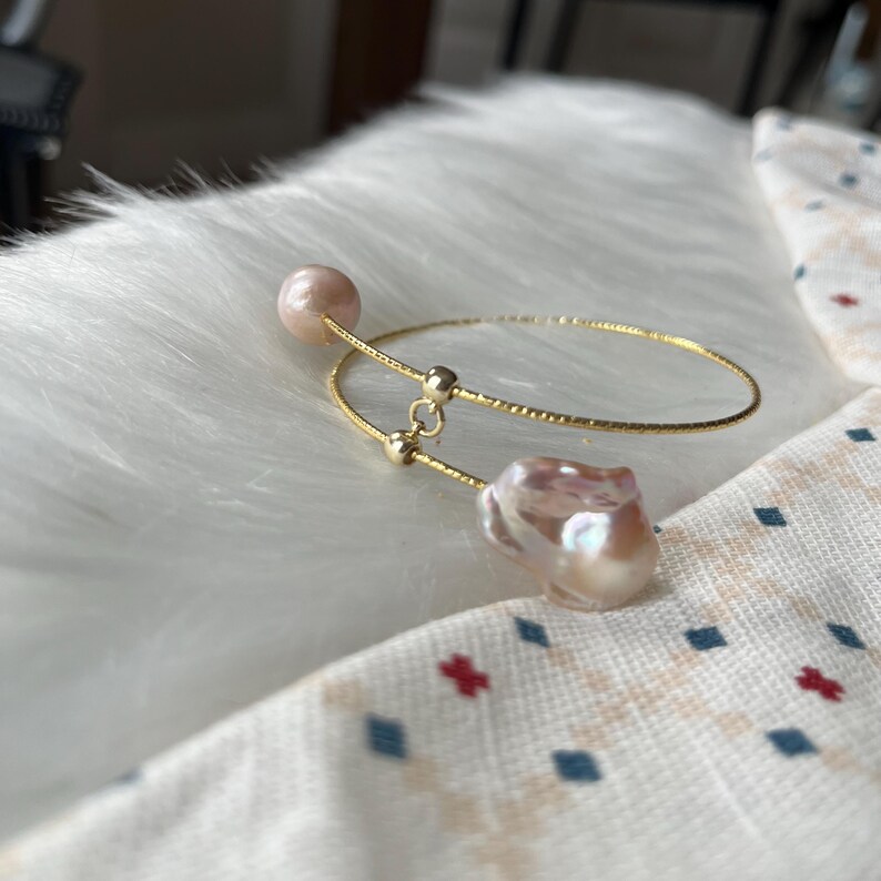 Peut inclure: Un bracelet jonc en or avec deux brins d&eacute;licats et un charme en perle. La perle est de forme baroque avec un &eacute;clat iridescent rose et blanc.