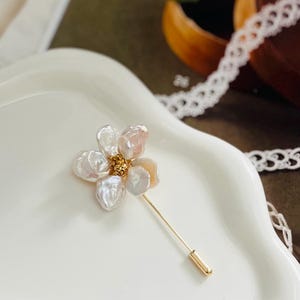 Puede incluir: Un delicado broche dorado con un dise&ntilde;o de flor de perla blanca y rosa. La flor tiene cinco p&eacute;talos y un centro dorado.