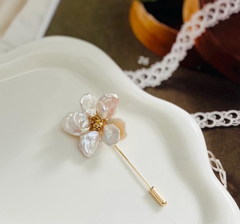 Puede incluir: Un delicado broche dorado con un dise&ntilde;o de flor de perla blanca y rosa. La flor tiene cinco p&eacute;talos y un centro dorado.