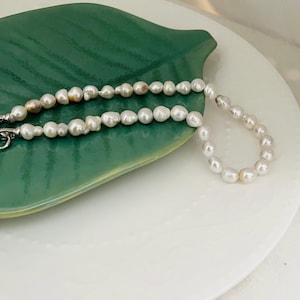 Puede incluir: Un collar de perlas blancas de forma irregular. El collar se muestra sobre un plato verde en forma de hoja, sobre un fondo blanco. Las perlas tienen un brillo lustroso.