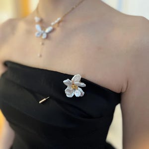 Puede incluir: Un broche de flor de perla blanca con detalles dorados. El broche est&aacute; prendido a un vestido negro sin tirantes.