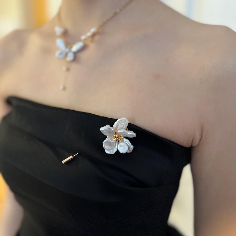 Puede incluir: Un broche de flor de perla blanca con detalles dorados. El broche est&aacute; prendido a un vestido negro sin tirantes.