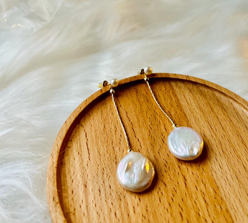 May include: A pair of gold dangle earrings with white pearl drops. The earrings are on a wooden surface.