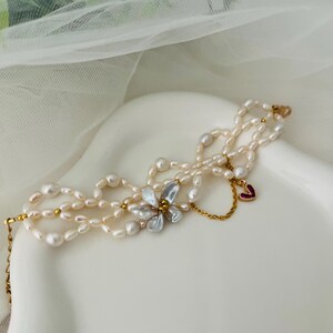 May include: A delicate pearl bracelet with multiple strands. The bracelet features a flower-shaped charm with iridescent petals and a small red heart charm. The pearls are off-white and the metal accents are gold-toned.