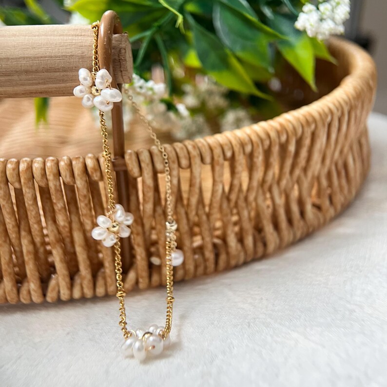 May include: A delicate gold chain bracelet with small white pearl flower accents. The bracelet is displayed on a woven wicker tray.