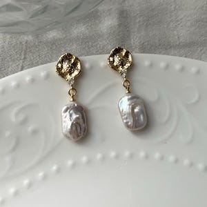 Puede incluir: Un par de pendientes de oro con un disco texturizado irregular y una perla barroca colgante. Cada pendiente tiene un peque&ntilde;o cristal transparente.