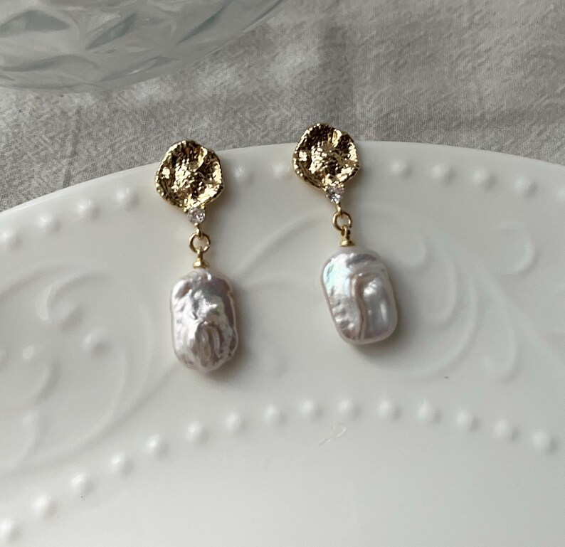 Puede incluir: Un par de pendientes de oro con un disco texturizado irregular y una perla barroca colgante. Cada pendiente tiene un peque&ntilde;o cristal transparente.