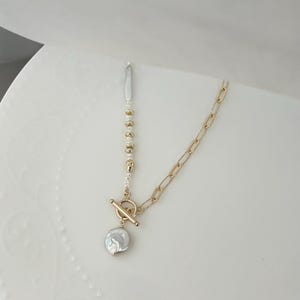 May include: A gold-coloured necklace with a toggle clasp and a large, irregular-shaped pearl pendant. The necklace features a chain link design on one side and a strand of small pearls and gold beads on the other.