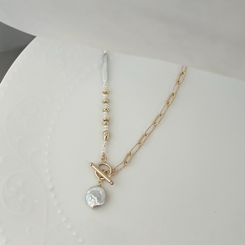 May include: A gold-coloured necklace with a toggle clasp and a large, irregular-shaped pearl pendant. The necklace features a chain link design on one side and a strand of small pearls and gold beads on the other.