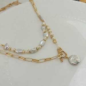 May include: A layered necklace with a gold chain and a strand of white baroque pearls. The necklace has a gold toggle clasp and a large, flat, white pearl pendant. The jewelry is displayed on a white surface.