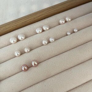 Peut inclure: Diverses boucles d'oreilles &agrave; clous en perles de diff&eacute;rentes tailles et couleurs, dont blanc et rose. Les boucles d'oreilles sont dispos&eacute;es sur une surface beige clair textur&eacute;e, avec une bordure en bois en haut.