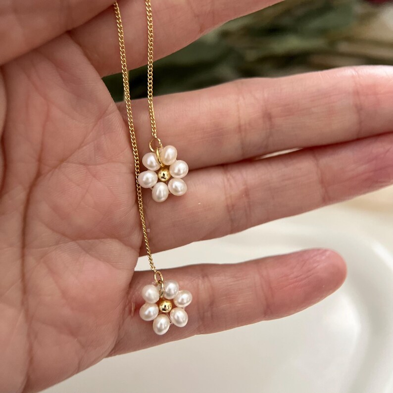 May include: A delicate gold chain necklace with two pearl flower pendants. Each flower is made of small white pearls arranged in a circular shape.