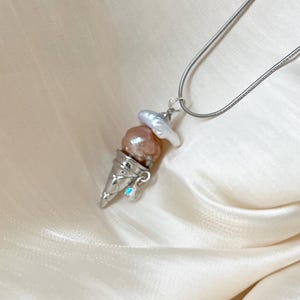 Peut inclure: Un collier en cha&icirc;ne argent avec un pendentif en forme de cornet de glace. Le cornet est rempli de trois perles roses et blanches.