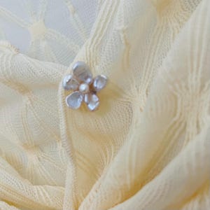 May include: A close-up of a pale yellow knitted fabric with a floral brooch. The brooch is silver-toned with a pearl center and petal-shaped accents. The fabric has a textured, open weave pattern.