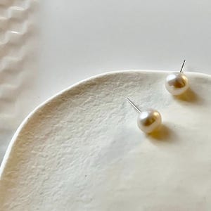 Peut inclure: Deux boucles d'oreilles en perles avec des tiges en argent reposent sur une surface blanche textur&eacute;e. Les perles sont de couleur cr&egrave;me et ont une forme lisse et ronde. Les boucles d'oreilles sont un bijou classique.