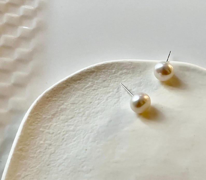 Peut inclure: Deux boucles d'oreilles en perles avec des tiges en argent reposent sur une surface blanche textur&eacute;e. Les perles sont de couleur cr&egrave;me et ont une forme lisse et ronde. Les boucles d'oreilles sont un bijou classique.