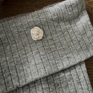 Może przedstawiać: Mała, kremowa ozdoba w kształcie róży spoczywa na złożonej sekcji szarej, prążkowanej tkaniny. Tkanina ma teksturowany wygląd. Róża jest szczegółowa z wieloma płatkami.