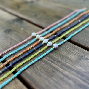 May include: A set of six necklaces with different colored beads and a pearl accent. The necklaces are arranged in a row on a wooden surface.