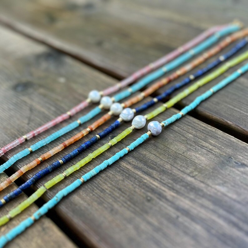 May include: A set of six necklaces with different colored beads and a pearl accent. The necklaces are arranged in a row on a wooden surface.