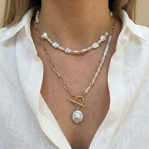 May include: Two-strand necklace featuring freshwater pearls and gold-tone chains. The top strand has irregular pearls, while the lower strand has a large, baroque pearl pendant. The necklace is worn against a white shirt.