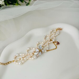 May include: A delicate pearl bracelet with a gold chain and clasp. The bracelet features multiple strands of white and cream-colored pearls, accented with gold beads and a small charm. A cluster of light blue and white petals adds a floral touch.